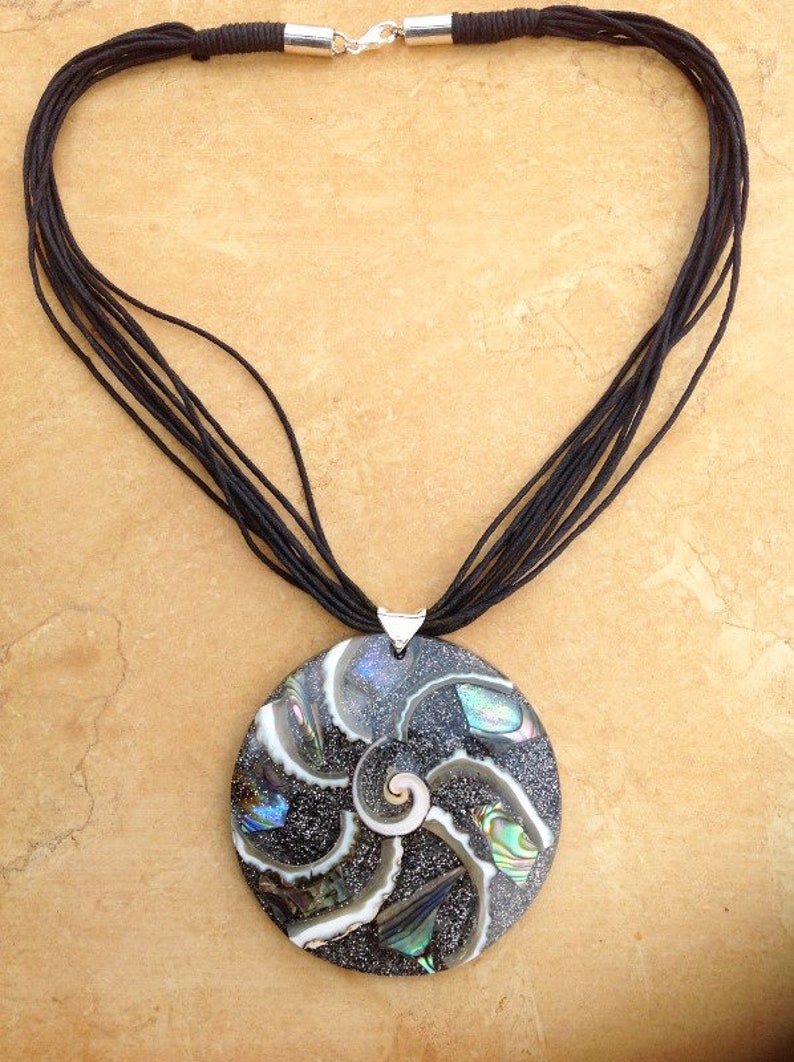 K&ouml;nnte beinhalten: Eine schwarze Schnurkette mit einem runden Anh&auml;nger, der ein Spiraldesign aus irisierendem Abalone-Muschel zeigt.