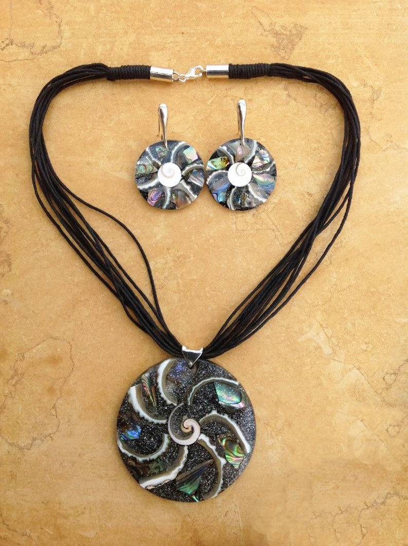 K&ouml;nnte beinhalten: Eine schwarze Halskette mit einem gro&szlig;en runden Anh&auml;nger aus schillerndem Abalone-Muschelschalen. Der Anh&auml;nger zeigt ein Spiralmuster. Die Halskette wird mit passenden Ohrringen mit kleineren runden Abalone-Muschelschalen-Anh&auml;ngern geliefert.