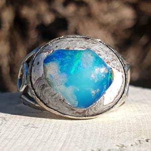 May include: A silver ring featuring a large, irregular, raw opal gemstone as the centrepiece. The opal displays vibrant flashes of bright blue and green colour. The metal setting has a textured, organic, and weathered appearance surrounding the stone.