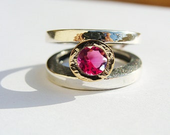 Könnte beinhalten: Ein silberner Ring mit einer goldenen Fassung, die einen großen, runden, rosa Edelstein hält. Der Ring hat ein einzigartiges Doppelband-Design, wobei der Edelstein prominent in der Mitte zur Geltung kommt. Auf hellem Hintergrund.