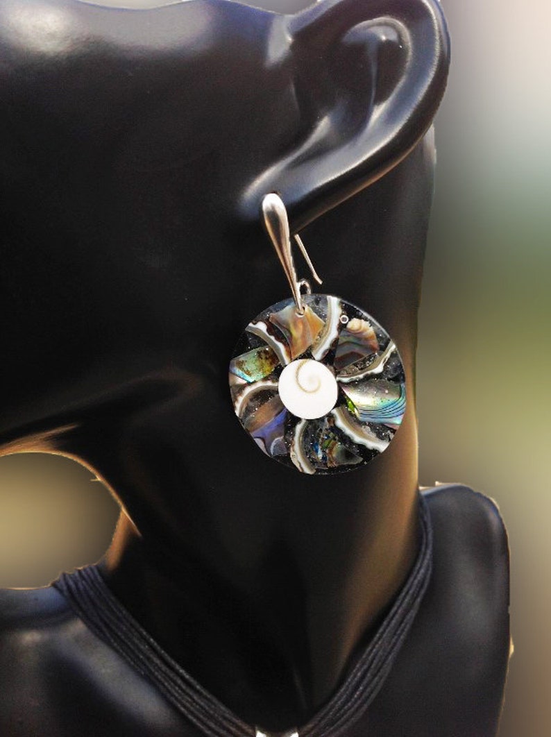 K&ouml;nnte beinhalten: Ein Paar silberne Ohrringe mit einem runden, schillernden Abalone-Muschel-Design. Die Muschel besteht aus mehreren Teilen von Abalone-Muschel, die einen Mosaik-Effekt erzeugen. Die Mitte der Muschel zeigt eine wei&szlig;e, spiralf&ouml;rmige Muschel.