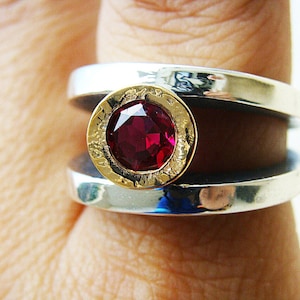 May include: A silver and gold ring with a red gemstone set in a circular bezel. The ring has a textured band and a wide, flat design.