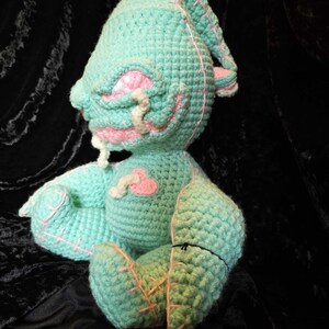 May include: A crocheted plush toy in the shape of a monster with a green body, pink accents, and a stitched-on heart. The toy has a stitched-on mouth and eyes.
