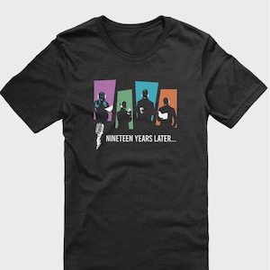 May include: A dark heather grey t-shirt with a graphic of five figures in silhouette, standing in front of colorful rectangular blocks. The text "NINETEEN YEARS LATER..." is printed below the graphic.
