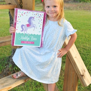 May include: A young girl holds a personalized binder with a unicorn design. The binder is pink with glitter trim and features the name "Madelyn Reece" and "First Grade". She wears a light blue dress and silver sandals.