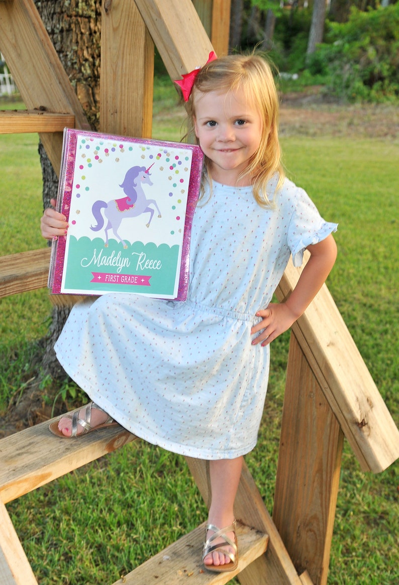 May include: A young girl holds a personalized binder with a unicorn design. The binder is pink with glitter trim and features the name "Madelyn Reece" and "First Grade". She wears a light blue dress and silver sandals.