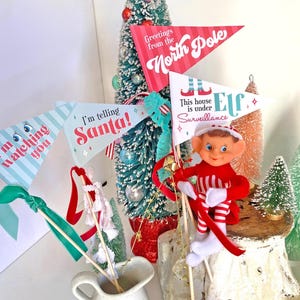 Peut inclure: Quatre drapeaux de Noël festifs avec des messages différents : "Je te surveille", "Je le dis à Santa !", "Salutations du pôle Nord" et "Cette maison est sous surveillance d'un lutin". Un petit lutin en peluche tient un bâton rouge et blanc rayé.