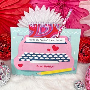 May include: A pink typewriter card with a message that says "You're the "write" friend for me. From: Madelyn". The card is surrounded by red and pink tissue paper.