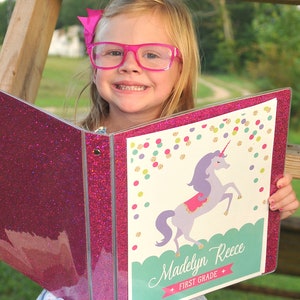 May include: A pink glitter binder with a unicorn design. The binder has a white background with a unicorn illustration, colorful confetti, and the name "Madelyn Reece" with "First Grade" below. The binder is open, revealing the inside.