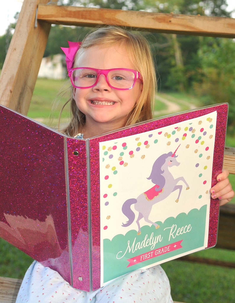 May include: A pink glitter binder with a unicorn design. The binder has a white background with a unicorn illustration, colorful confetti, and the name "Madelyn Reece" with "First Grade" below. The binder is open, revealing the inside.