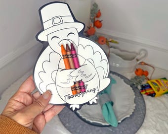 Puede incluir: Un dibujo para colorear con forma de pavo blanco y negro con un sombrero de peregrino, que sostiene varios crayones. El texto "Thanksgiving!" está impreso en el pavo. Los crayones son de color naranja, marrón y rosa.