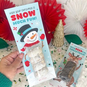 Op de afbeelding: Een lichtblauwe kerstcadeaulabel met een sneeuwpopillustratie en de tekst "Hope your Christmas is SNOW MUCH FUN!". Het label bevat een doorzichtige zak met koekjes. Een tweede cadeaulabel met een rendierillustratie is ook zichtbaar.
