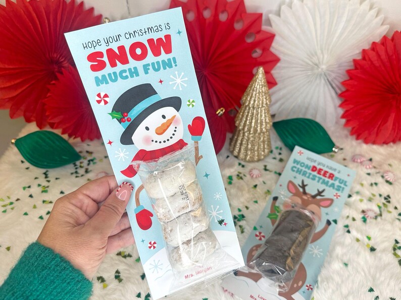 Op de afbeelding: Een lichtblauwe kerstcadeaulabel met een sneeuwpopillustratie en de tekst "Hope your Christmas is SNOW MUCH FUN!". Het label bevat een doorzichtige zak met koekjes. Een tweede cadeaulabel met een rendierillustratie is ook zichtbaar.