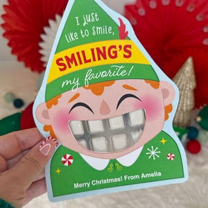 May include: A Christmas-themed card shaped like an elf's head, with the text "I just like to smile, Smiling's my favorite!" and "Merry Christmas! From Amelia." The card features a clear window revealing a pack of white gum.