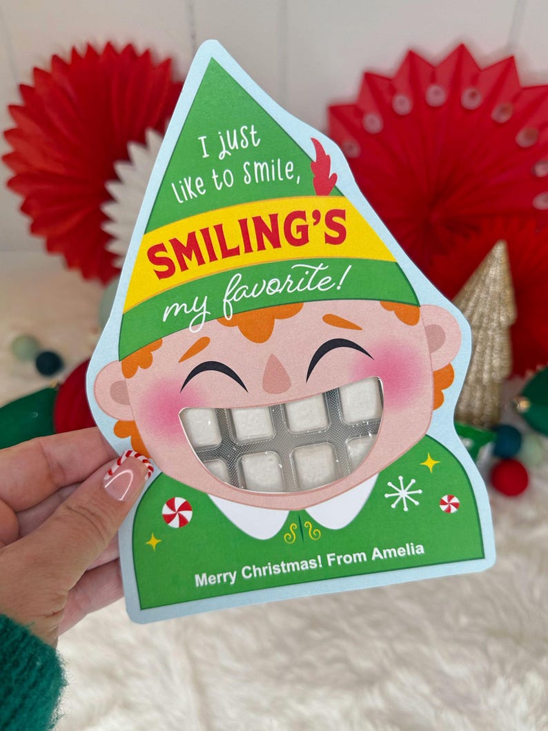 May include: A Christmas-themed card shaped like an elf's head, with the text "I just like to smile, Smiling's my favorite!" and "Merry Christmas! From Amelia." The card features a clear window revealing a pack of white gum.