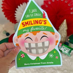 May include: A Christmas-themed card shaped like an elf with the text "I just like to smile, Smiling's my favorite!" and "Merry Christmas! From Amelia." The card holds a pack of gum.