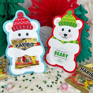 Könnte beinhalten: Zwei Geschenkhalter in Form von Eisbären, einer blau und einer rot, die jeweils Haribo-Gummibonbons enthalten. Der blaue Bär trägt einen roten Hut, und der rote Bär hat einen grünen Hut und Schal. Auf dem roten Bären steht "Wishing you a BEARY Merry Christmas!"