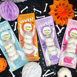 以下が含まれることがあります： 「A Yummy Mummy for You to Unwrap」のテキストが入った、ハロウィーンをテーマにした4つのカラフルなお菓子パッケージ。各パッケージにはドーナツが詰まっています。装飾には、ペーパーハニカムボール、カボチャ、骨型のキャンディーが含まれています。