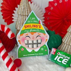 May include: A festive Christmas-themed display featuring a smiling elf-shaped box with gum, the text "I just like to smile, Smiling's my favorite!" and "Merry Christmas! From Amelia". Includes a box of Dentyne Ice gum, red and gold decorations.