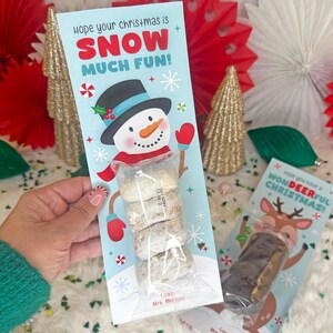 Op de afbeelding: Een lichtblauw kerstcadeaulabel met een sneeuwpopafbeelding en de tekst "Hope your christmas is SNOW MUCH FUN!". Het label bevat een doorzichtige zak met koekjes. Een tweede label toont een rendier.