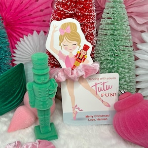 Puede incluir: Una figurita de cascanueces de terciopelo verde se encuentra junto a un adorno de terciopelo rosa y una tarjeta rosa y blanca con el texto "Dancing with you is Tutu FUN! Merry Christmas! Love, Hannah".