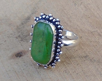 Puede incluir: Un anillo de plata con una piedra preciosa verde engastada en un engaste decorativo. El engaste tiene un borde festoneado con pequeñas cuentas.