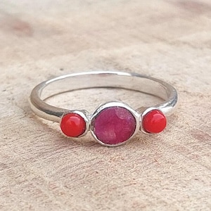 May include: A silver ring featuring a large, round, ruby-colored center stone, flanked by two smaller, bright red stones. The ring is set against a light-colored, textured surface.
