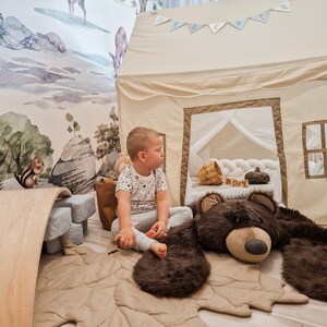 May include: A child sits on a brown leaf-shaped rug with a large brown teddy bear. The rug is in front of a beige play tent with a white mesh window. The tent has a blue and white banner that says "Happy" hanging above it.