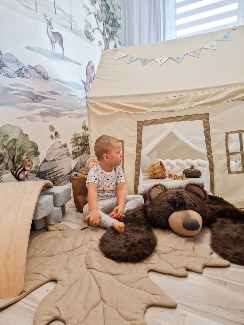 May include: A child sits on a brown leaf-shaped rug with a large brown teddy bear. The rug is in front of a beige play tent with a white mesh window. The tent has a blue and white banner that says "Happy" hanging above it.