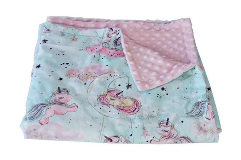 May include: A soft, plush baby blanket with a light blue background featuring a unicorn design. The blanket has a pink minky dot fabric on the reverse side.