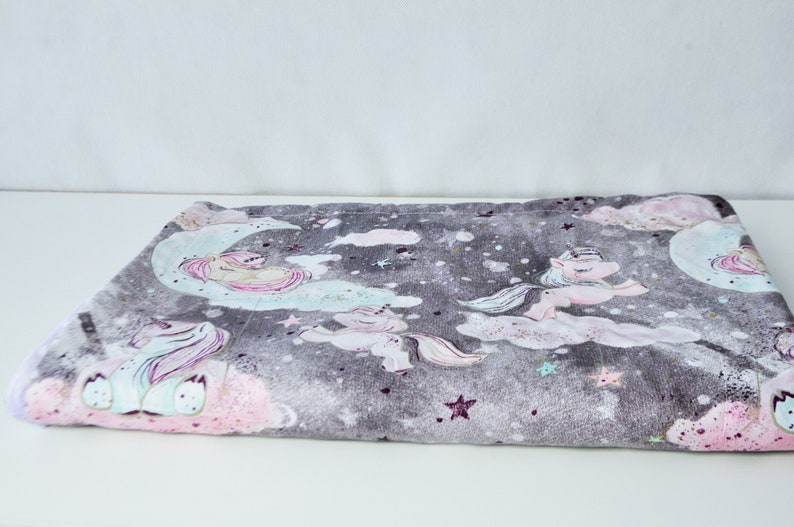 May include: A soft grey and pink baby blanket with a unicorn and moon design. The blanket features a repeating pattern of unicorns, moons, and stars.