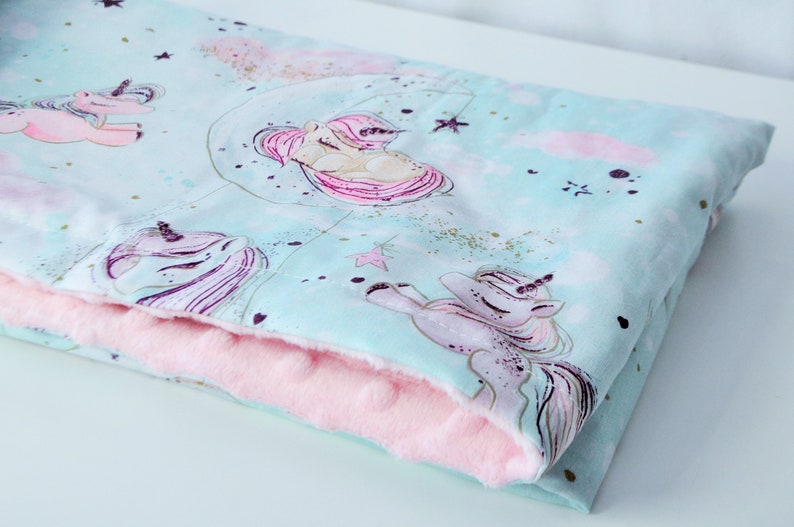 May include: A soft, light blue and pink baby blanket with a unicorn print. The blanket is made of two layers of fabric, with a soft, plush pink layer on the inside and a light blue layer with a unicorn print on the outside.