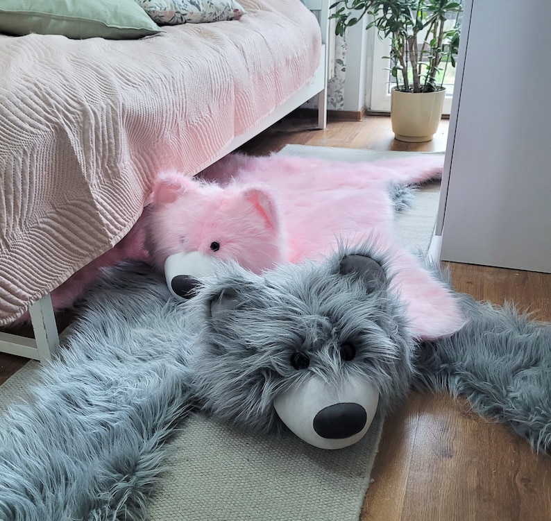 May include: A gray and pink faux fur rug with a gray wolf and a pink bear plush toy. The rug is on a wooden floor.