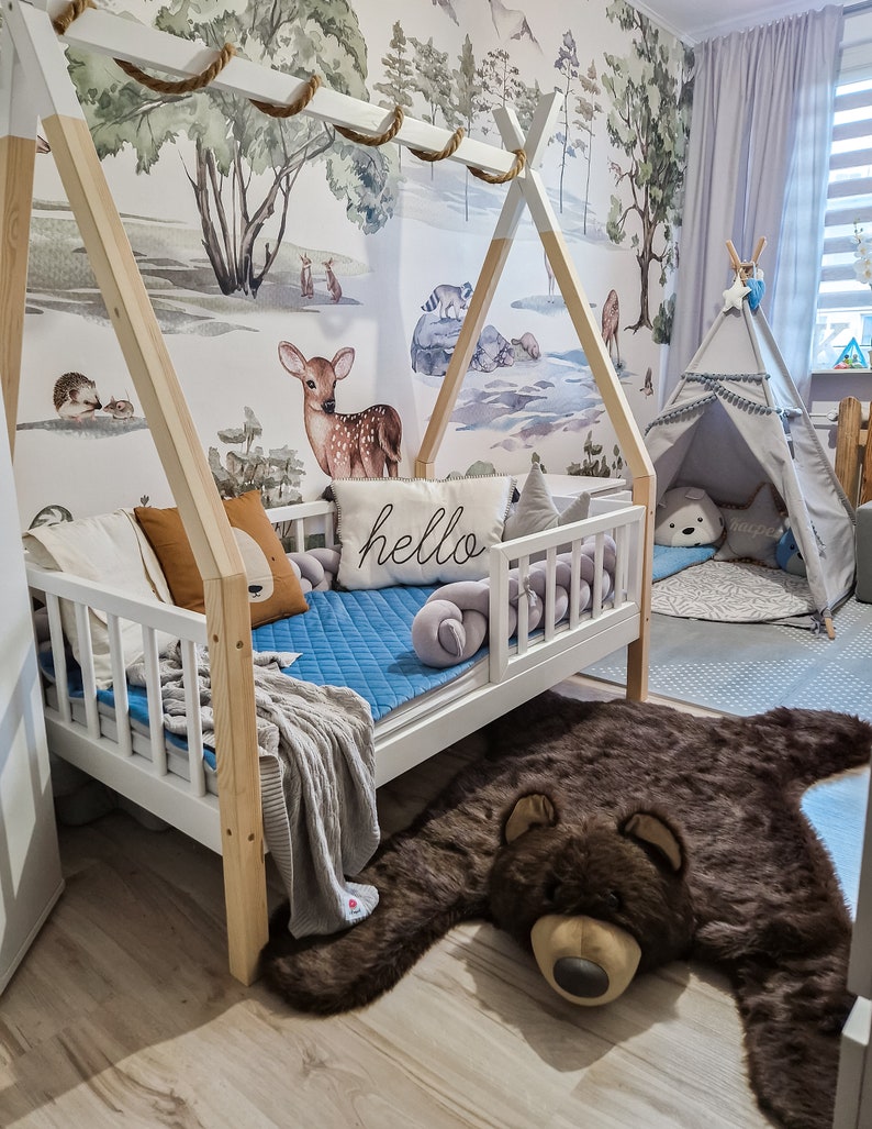 May include: A white wooden teepee bed frame with a blue and white quilted mattress and a brown bear rug on the floor. The teepee bed frame has a rope and wooden dowel at the top. The wall behind the bed has a watercolor mural of a forest scene with animals.