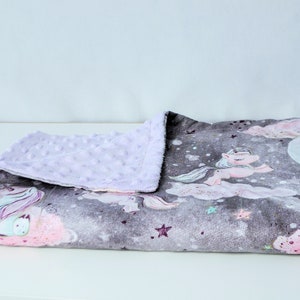 May include: A soft, plush baby blanket with a grey and white background featuring a unicorn and stars. One side of the blanket is a soft, lavender minky fabric.