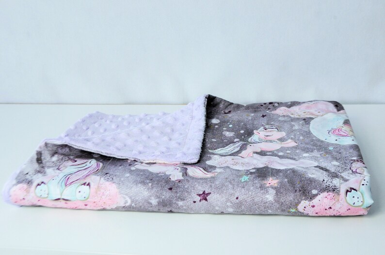 May include: A soft, plush baby blanket with a grey and white background featuring a unicorn and stars. One side of the blanket is a soft, lavender minky fabric.