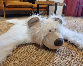 May include: A white faux fur polar bear rug with a brown nose and black eyes. The rug is lying on a woven rug.