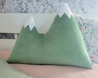 May include: A green velvet mountain-shaped pillow with white felt peaks. The pillow is on a pink bedspread.