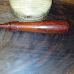 May include: A close-up of a wooden mallet with a reddish-brown finish. The mallet has a rounded head and a handle with a dark brown band.