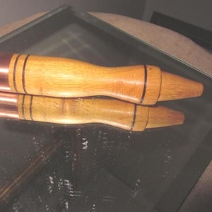 Puede incluir: Dos tapones de botella de madera con detalles de cobre y rayas negras. Los tapones están hechos de madera clara y tienen un diseño cónico. Se muestran sobre una superficie reflectante.