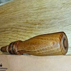May include: A wooden bottle stopper with a brown and natural wood grain finish. The stopper has a rounded end and a slightly tapered shape.