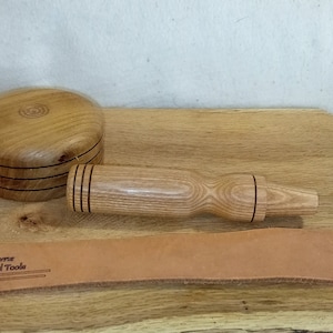 Puede incluir: Un mazo de madera con una cabeza redonda y un mango. El mazo está hecho de madera marrón claro con rayas marrones oscuras. El mazo está sobre una superficie de madera con una correa de cuero que dice "Vallone Wood Tools".