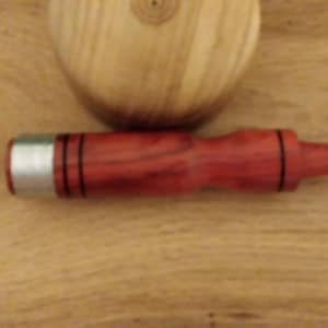 Puede incluir: Un tapón de botella rojo y plateado con líneas negras. El tapón está hecho de madera y metal. Una esfera de madera está en el fondo.