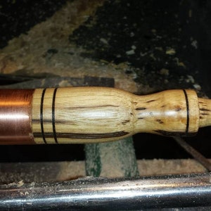 Puede incluir: Un mango de madera con una banda de cobre y detalles negros se está girando en un torno. El mango está hecho de madera clara y oscura, creando un patrón a rayas.