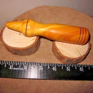 Puede incluir: Un tapón de botella de madera artesanal con acabado marrón claro, apoyado sobre dos pequeñas rodajas de madera. El tapón tiene un cuerpo redondeado con tres ranuras marrones oscuras y mide unos 15 cm de largo.