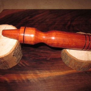 Puede incluir: Un tapón de botella de madera hecho a mano con un pico cónico y un cuerpo bulboso. El tapón es de madera de color marrón rojizo, con tres bandas negras cerca de la base. Descansa sobre dos pequeñas rodajas de madera redondas.