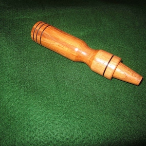 Puede incluir: Un reclamo de pato de madera con un acabado marrón claro. El reclamo tiene un cuerpo cilíndrico con tres anillos negros cerca de la parte superior y una boquilla acampanada. El reclamo está sobre una superficie verde.