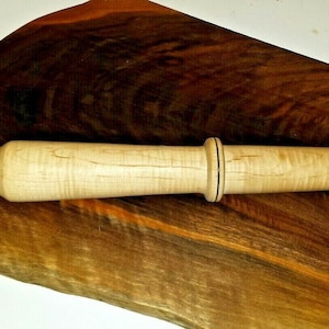 Puede incluir: Un rodillo de madera con un acabado marrón claro y detalles negros. El rodillo está apoyado sobre una superficie de madera marrón oscuro.