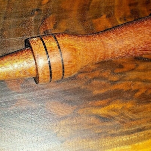 Puede incluir: Una clavija de madera con un extremo cónico y anillos negros alrededor del centro. La clavija está hecha de madera marrón rojiza.