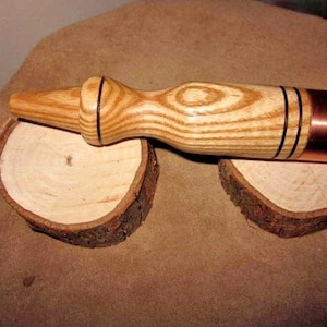 May include: A wooden mallet with a copper end, resting on a wooden surface. The mallet has a natural wood grain pattern and black accents.